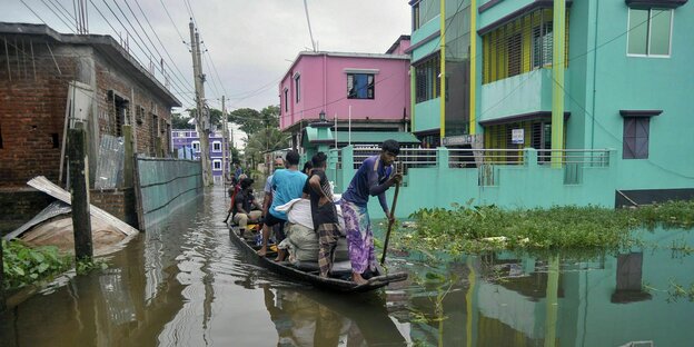 Mehrere Menschen staken sich auf einem flachen Boot durch überflutete Straßen mit bunten Häusern
