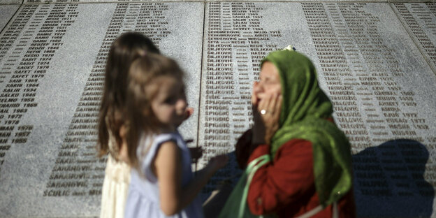 Eine ältere Frau und zwei Mädchen stehen vor einer Gedenktafel in Srebrenica