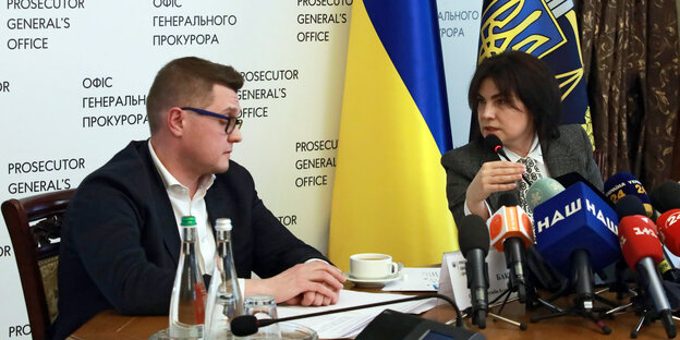 Ein mann und eine Frau sitzen vor ukrainischer Flagge an einem Tisch mit vielen Mikrofonen