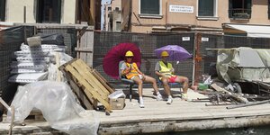 Ein Performancekünstlerpaar sitzt mit Schirmen und Arbeitswesten vor einer Baustelle in einer italienischen Stadt