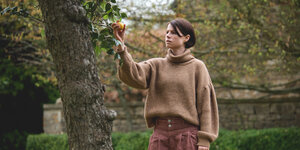 Jessie Buckley steht im Garten und pflückt einen Apfel von einem Baum.