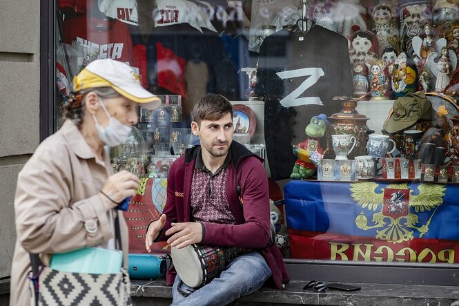 Ein Straßenmusiker sitzt vor einem Laden, eine alte Frau geht an ihm vorbei