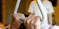 Eine Pflegehausbewohnerin eines Seniorenzentrums hält sich in ihrem Bett an einem Haltegriff fest, im Hintergrund steht eine Pflegekraft.