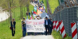 Demonstrierende Menschen gehen an einem Zaun entang, sie tragen ein Plakat mit der Aufschrift "Stoppt den Krieg. Frieden für die Ukraine und ganz Europa"