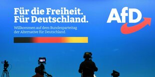 Blaues AFD-Plakat, davor stehen in größerem Abstand Kameraleute