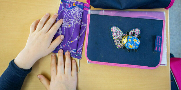 A cloth mask and a pencil case lie on a school desk.  A child's hands are on the mask.
