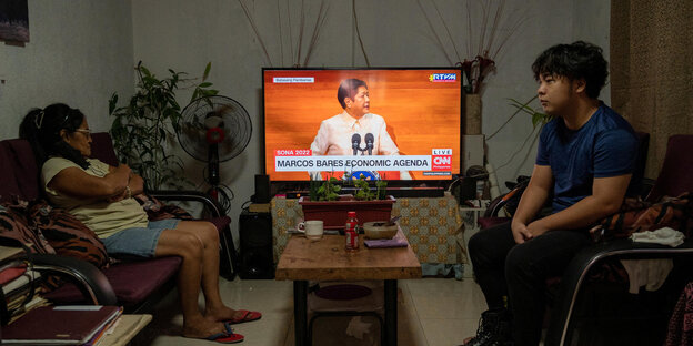In einem Wohnzimmer verfolgen zwei Menschen die auf einem Sofa sitzen die Antrittsrede des Präsidenten im Fernsehen.