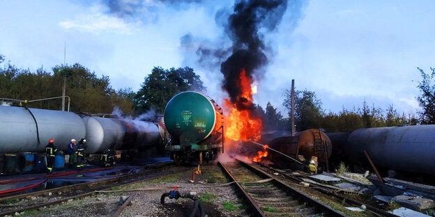 A burning tank car, with black smoke rising above it, with firefighters extinguishing it