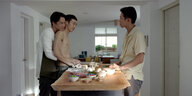 Drei junge Männer stehen an einer Küchentheke, einer umarmt den anderen von hinten