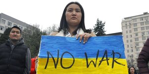 Eine Demonstrantin trägt ein blau-gelbes Plakat mit der Aufschrift "No War"