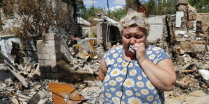 Eine Frau weint vor einem zerstörten Haus. Sie trägt eine Bluse mit Margeritenblumen