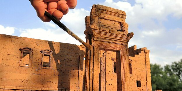Eine Hand mit Pinsel streift das Korkmodell des Baaltempel von Palmyra