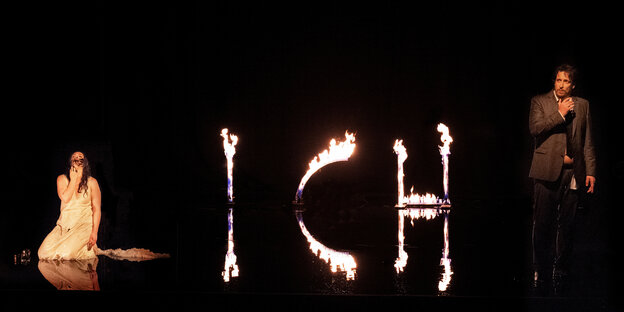 Links sitzt eine Frau auf einer dunklen Bühne, rechts steht ein Mann, zwischen ihnen zeichnen Flammen die Buchstaben i c h