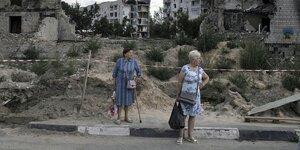 Zwei Frauen stehen vor Häuserruinen, sie tragen Einkaufstaschen