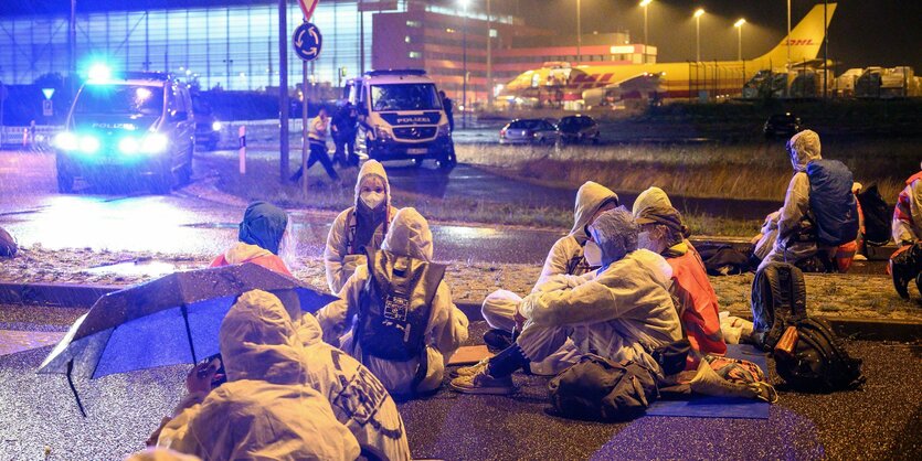 Aktivisten sitzen in Schutzanzügen vor einer Flughafeneinfahrt