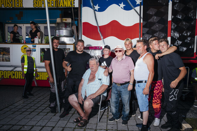 Vor einem Aufsteller mit US-Flagge stehen und sitzen neun männliche Mitglieder der Familie Frank. Links daneben stehen vor und hinter der Popcorn-Theke drei Frauen