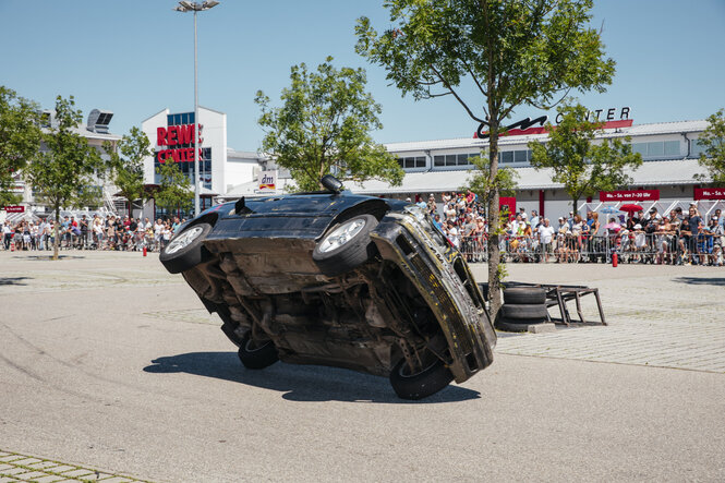 Ein dunkles Auto fährt auf zwei Rädern am Publikum vorbei. Im Hintergrund ist ein Rewe-Markt zu sehen