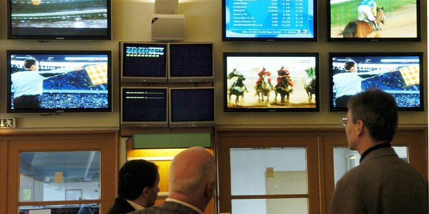 Drei Männer beobachten in einem Wettlokal die vielen Bildschirme, auf denen unter anderem Pferderennen und Quoten zu sehen sind