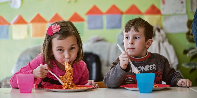 Ein Mädchen isst Spagetti mit dem Mund, ein Junge hält eine Gabel und guckt in den Raum