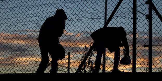 Zwei Flüchtlinge überqueren einen Zaun in der Dunkelheit.