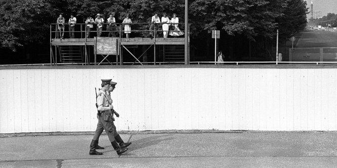 Zwei Grenzer laufen an der Berliner Mauer entlang. Im Hintergrund stehen Westberliner Touristen auf einer Brüstung und schauen auf die Grenzer rüber.