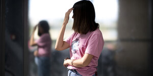 Eine Frau mit dunklen Haaren, die ihr Gesicht verdecken, steht im Profil mit rosa Shirt und Jeans