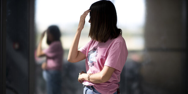 Eine Frau mit dunklen Haaren, die ihr Gesicht verdecken, steht im Profil mit rosa Shirt und Jeans