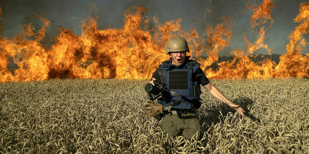 Ein Mann mit Schutzhelm und -bekleidung rennt durch ein Weizenfeld. Im Hintergrund: Flammen