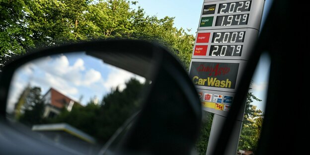 Blick aus dem Auto zeigt Tankstellen-Preise, durch den Seitenspiegel sieht man eine Wohnsiedlung.