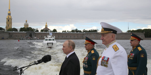Putin und Militärs stehen vor einem Mikrofon, im Hintergrund ein Schiff.