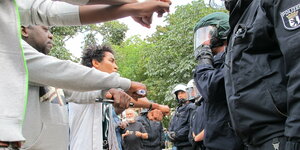 Demonstrierende stehen in einer Reihe gegenüber von Polizist:innen und halten die Hände überkreuz, als symbolische Aufforderung, verhaftet zu werden.