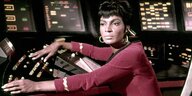 Nichelle Nichols in ihrere Rolle als Lieutenant Uhra in "Star Trek". Sie blickt Richtung Kamera, trägt ihre weinrote Uniform und riesige, goldene Ohrringe. Ihre Hände bewegen sich über das große Schaltpult auf der Brücke vor ihr.