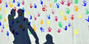 Drei Schatten zeichnen sich auf einer Wand voller Abdrücke von Kinderhänden ab.