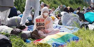 Demonstrantin im Plastikanzug zwischen anderen Menschen auf einer Wiese, in der Hand ein Schild "kein Grad mehr"