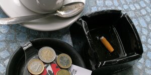 Detailaufnahme einer Kaffeetasse, eines Aschenbechers und eines Tellers auf dem Kleingeld liegt