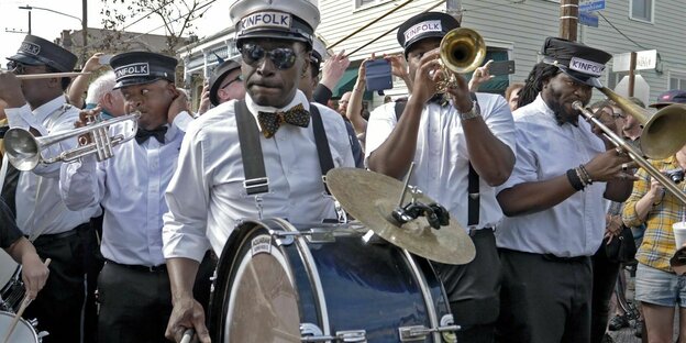 Eine Marching Band mit Trompeten und Trommeln spielt in New Orleans. Die Mitglieder tragen weiße Hemden und Mützen mit der Aufschrift "Kinfolk"