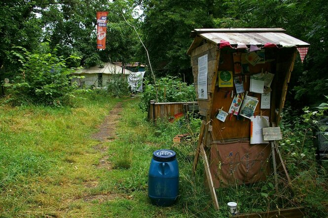 Verwilderter Garten mit einer Infotafel, an der viele Zettel und Broschüren befestigt sind