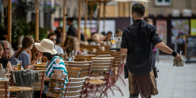 Eine Servicekraft läuft im Außenbereich eines Restaurants an Gästen vorbei.