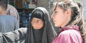 Eine ältere Frau im Tschador zeigt auf etwas außerhalb des Fotos, eine jüngere mit Zopf und Hemd blickt in die entsprechende Reichtung.