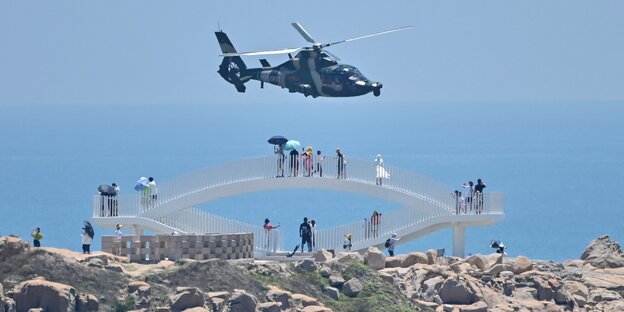Ein MIlitärhubschrauber über einer felsigen Küste und einer Brücke, worauf Menschen mit Sonnenschirmen stehen und Fotos machen