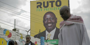 Passanten und Passantinnen gehen an einer Mauer entlang, im Hintergrund ein Wahlkampfplakat von William Ruto
