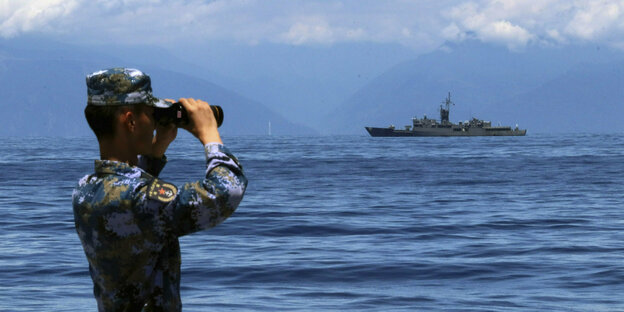 Ein Soldat schaut durch ein Fernglas auf ein Militärschiff im Meer