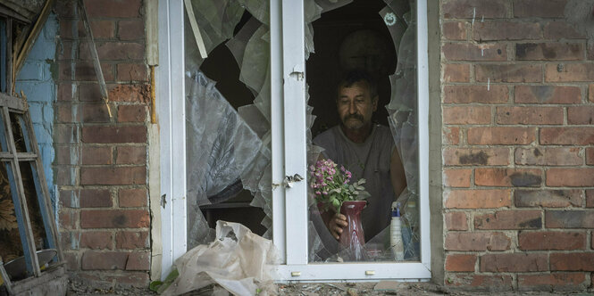 Ein Mann steht in einem Haus hinter einem durch Beschuss des russischen Militärs beschädigten Fenster und stellt eine Vase auf die Fensterbank