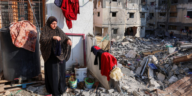 Eine Frau hängt Wäsche über eine Mauer, im Hintergrund zerstörte Häuser