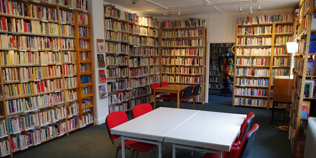 Ein Raum mit gefüllten Bücherregalen an den Wänden. Im Vordergrund steht ein Tisch mit vier roten Stühlen.