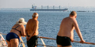 Zwei Männer und eine Frau in Badebekleidung betrachten ein Frachtschiff beim Auslaufen