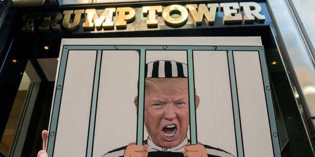 Auf einem Pappschild sieht man Trump hinter Gittern, im Hintergrund sieht man den Schriftzug des Trump-Towers