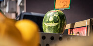 Ein Gemüsehändler hat an einem Verkaufsstand vor seinem Geschäft einer Wassermelone ein Gesicht geschnitzt.