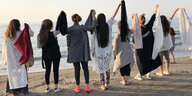 Eien Gruppe Frauen von hinten aufgenommen halten ihre Schleier an einem Strand in die Höhe in ihren rechten Händen