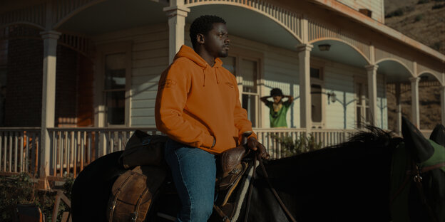 OJ sitzt auf einem Pferd vor der Haywood-Ranch, Emerald geht auf deren Veranda entlang.